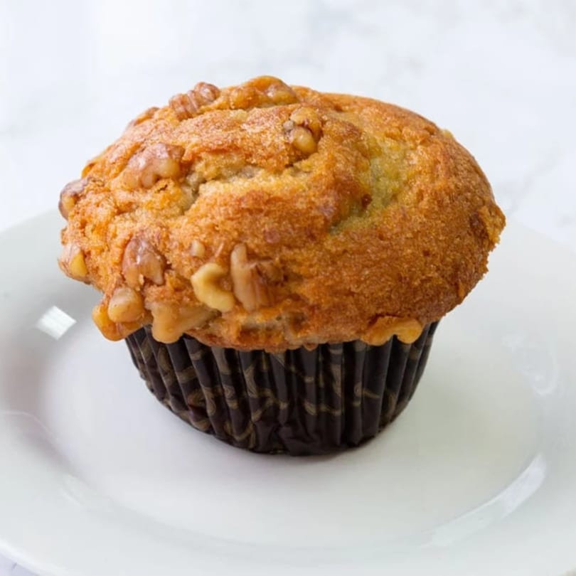 A golden-brown muffin with walnuts, in a dark brown wrapper, sits on a white plate.