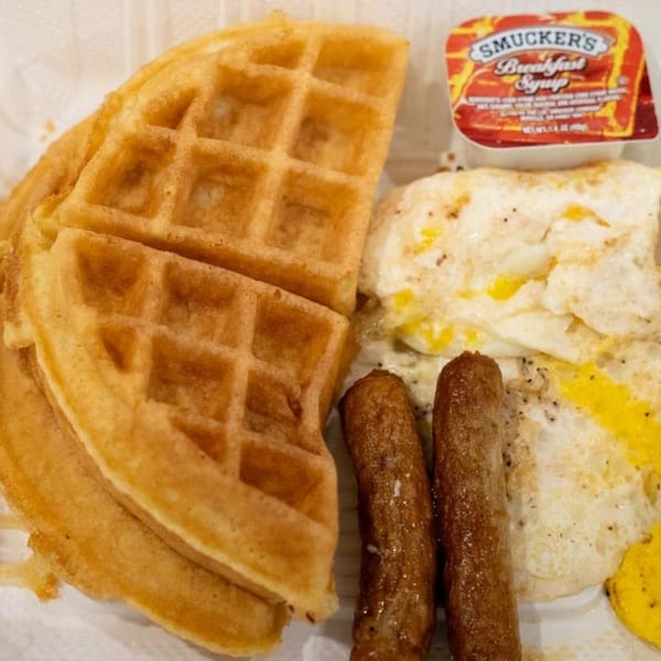 Waffles, fried eggs, sausage links, and a Smucker's syrup packet on a white plate.