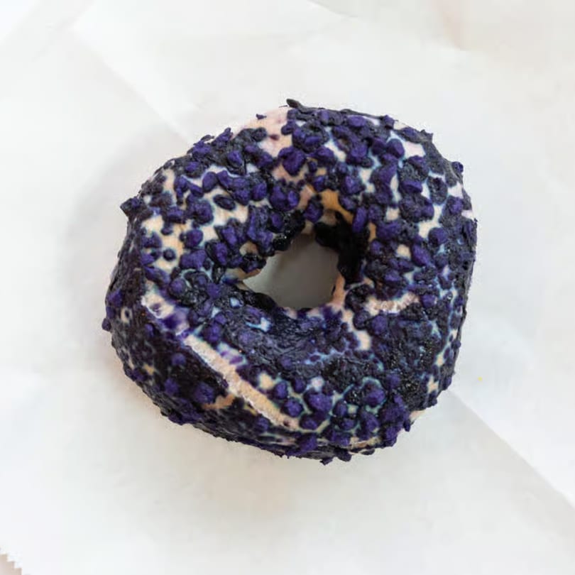 A Raceway Bagel with purple and black sprinkles on a white background.